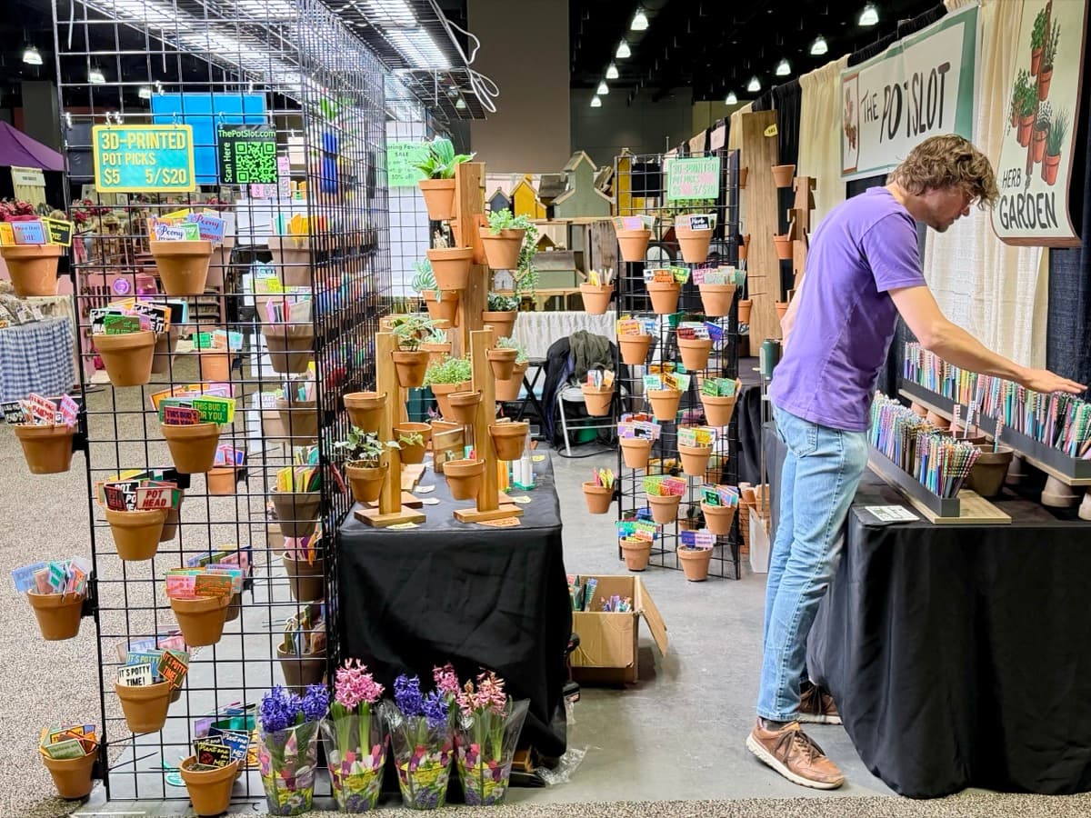 Will setting up The Pot Slot booth at an indoor craft show, organizing colorful 3D-printed garden picks and pot hangers