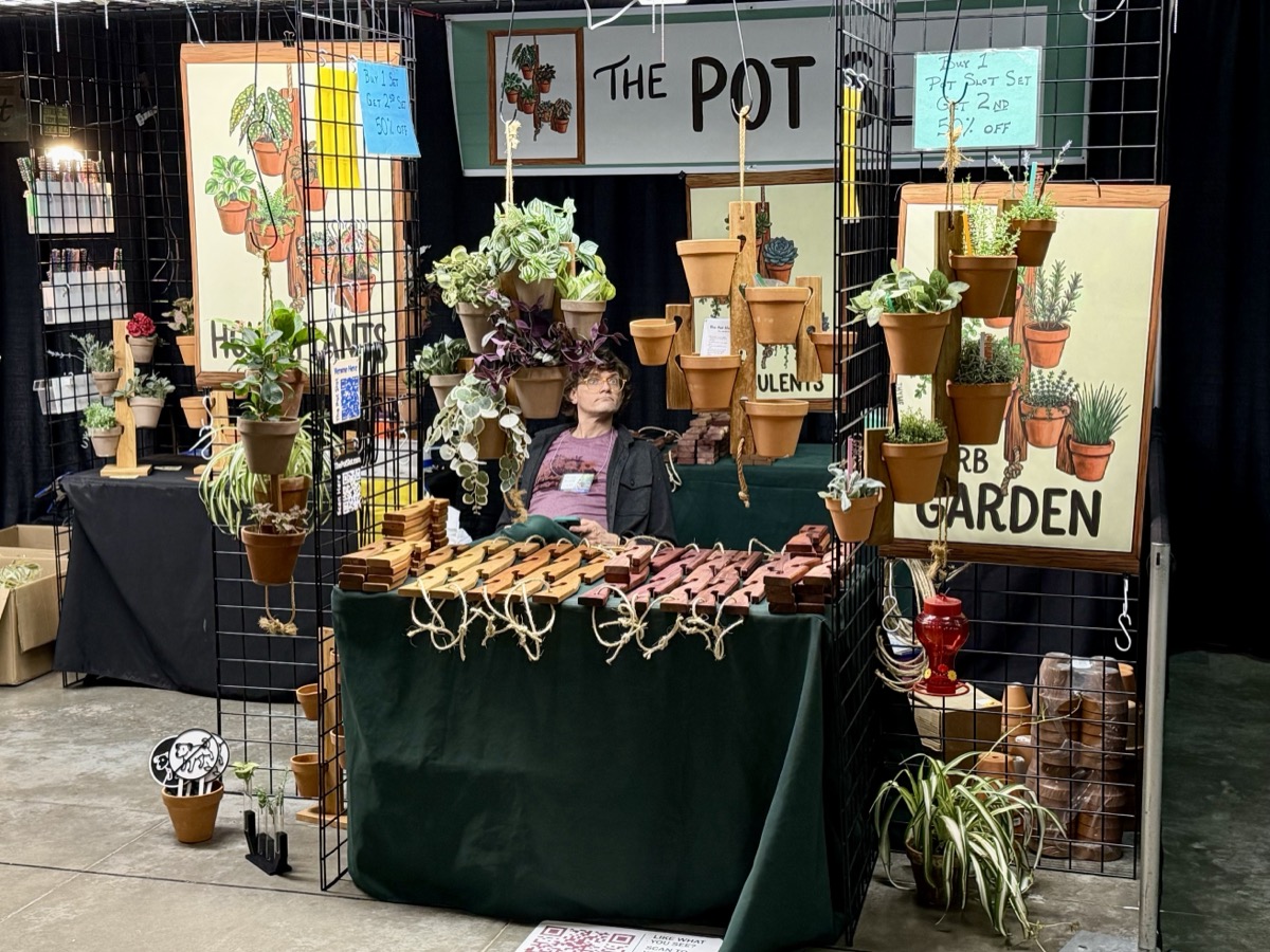 The Pot Slot booth at an indoor craft show with wooden pot hangers, terra cotta pots of succulents and houseplants, and hand-painted signs