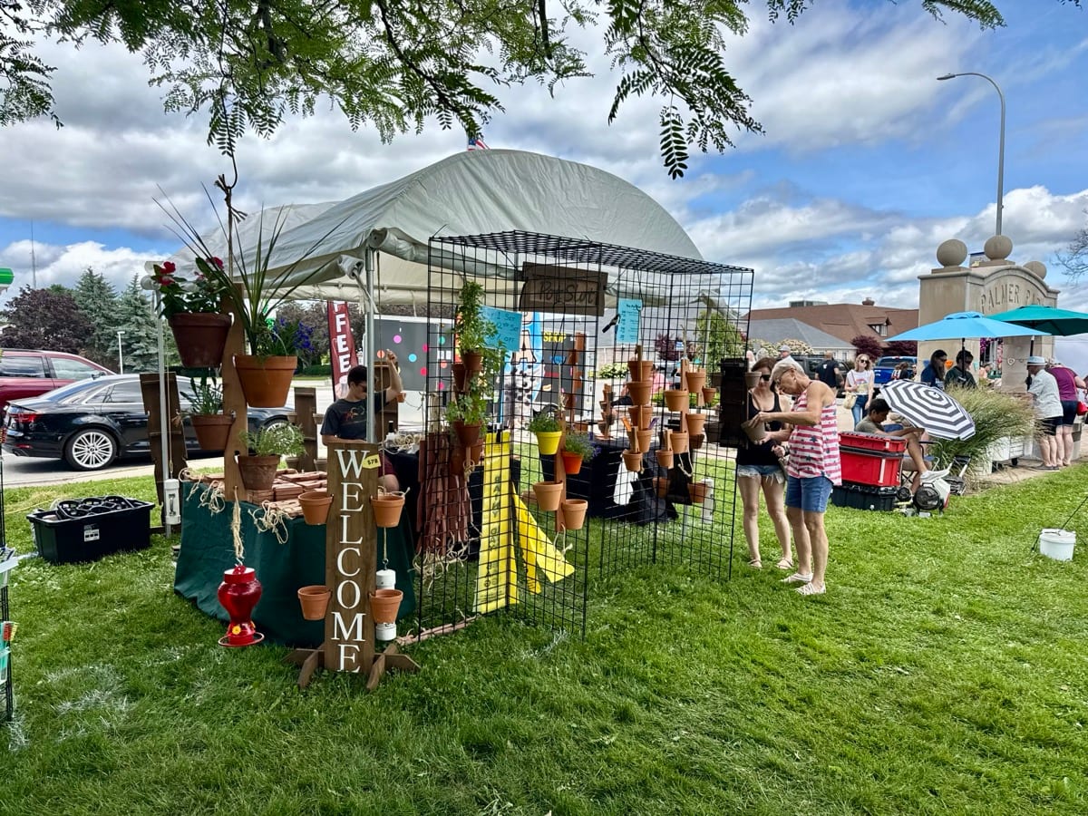 The Pot Slot booth at an outdoor farmers market with wooden pot hangers, terra cotta pots, and customers browsing on a sunny day